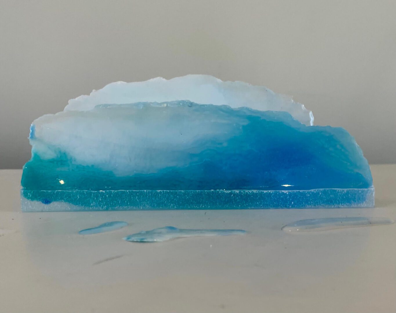 Available, July, 2025. A resin art piece resembling a cross-section of the ocean, with varying shades of blue, displayed on a white surface.