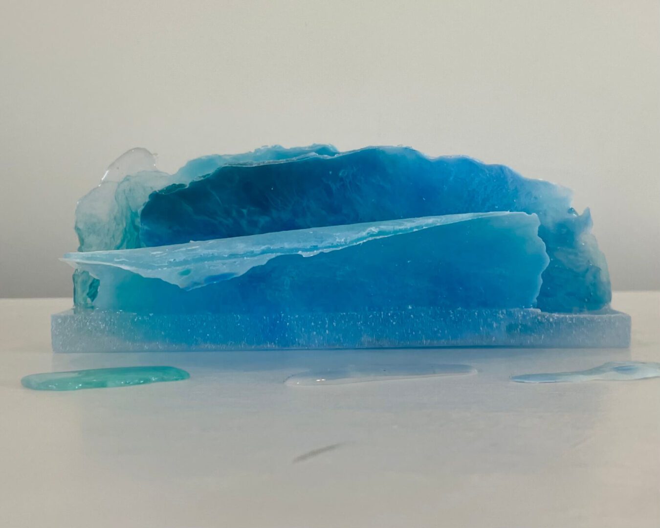 Available, July, 2025. A translucent blue resin sculpture resembling an ice formation, displayed on a white surface with droplets around it.